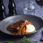 A plate of sliced steak with Essential Cuisine No1 Beef Gravy 1.5kg, garnished with herbs, next to mashed potatoes, accompanied by a glass of red wine and water, set on a dark table.