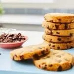 A stack of chocolate chip cookies on a kitchen counter with a bowl of Dr. Oetker Chocolate Chips Milk 750g and a blue napkin nearby.