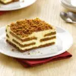 A slice of Lotus Biscoff Crumb 750g with multiple layers and a crumbly topping, served on a white plate with a red napkin on a wooden table.