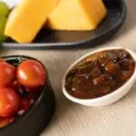 A small bowl of Arran Fine Foods Autumn Fruit Chutney 2.35kg next to fresh tomatoes and a plate of bread with grapes in the background.