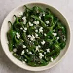 Charred Padron peppers with feta crumble and lemon zest created by Nicol Retailer, served in a rustic ceramic bowl and drizzled with olive oil.