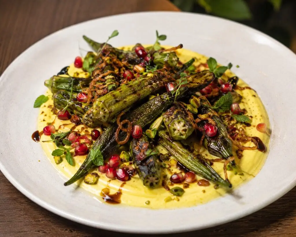 Charred fresh okra served with saffron yoghurt, crispy shallots, pistachios, pomegranate seeds, and pomegranate molasses on a gourmet fine-dining plate – recipe and image created by Nicol Retailer Limited.