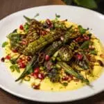 Charred fresh okra served with saffron yoghurt, crispy shallots, pistachios, pomegranate seeds, and pomegranate molasses on a gourmet fine-dining plate – recipe and image created by Nicol Retailer Limited.