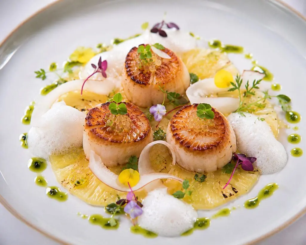Michelin-star seared king scallops served with fresh coconut foam, pineapple carpaccio, citrus herb oil, finger lime pearls, coconut ribbons, micro herbs, and edible flowers on an elegant fine-dining plate – recipe and image created by Nicol Retailer Limited.
