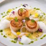 Michelin-star seared king scallops served with fresh coconut foam, pineapple carpaccio, citrus herb oil, finger lime pearls, coconut ribbons, micro herbs, and edible flowers on an elegant fine-dining plate – recipe and image created by Nicol Retailer Limited.