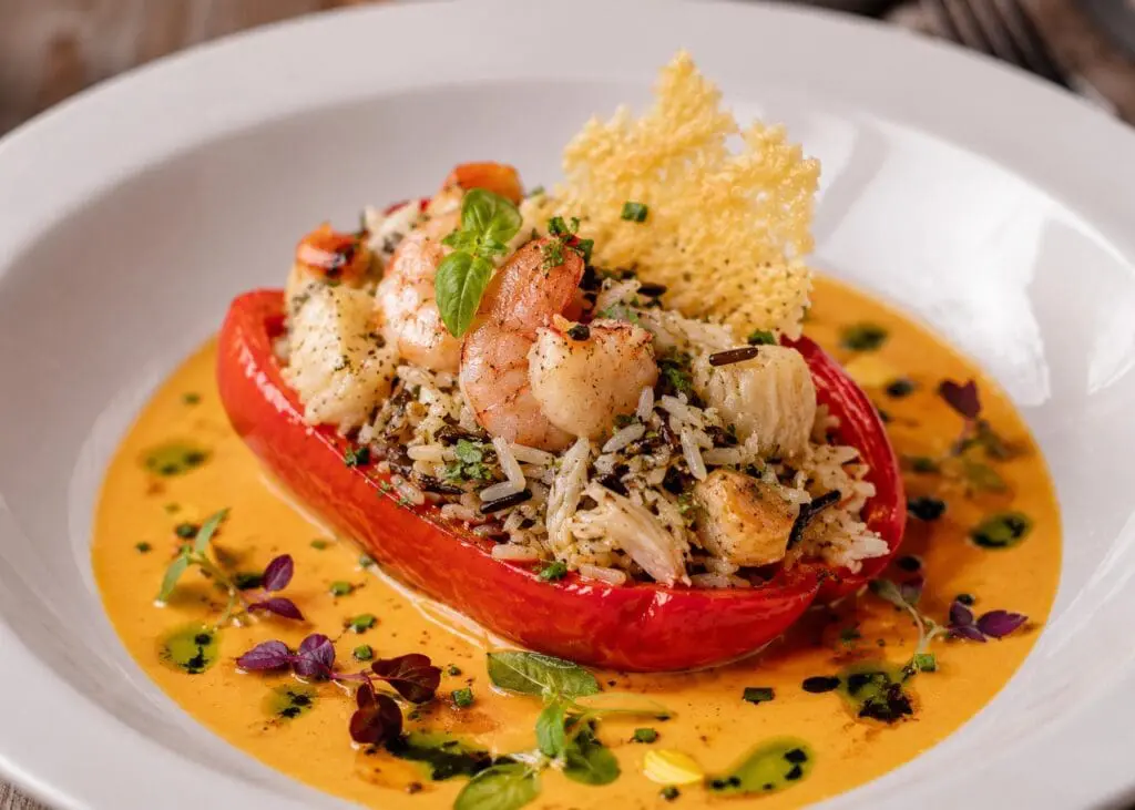 Michelin-star Mediterranean seafood stuffed Romano pepper filled with Tilda Easy Cook Basmati & Wild Rice, king prawns, scallops, and crab, served with saffron shellfish bisque, Parmesan tuile, herb oil, edible flowers, and micro herbs on a fine-dining white plate.