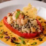 Michelin-star Mediterranean seafood stuffed Romano pepper filled with Tilda Easy Cook Basmati & Wild Rice, king prawns, scallops, and crab, served with saffron shellfish bisque, Parmesan tuile, herb oil, edible flowers, and micro herbs on a fine-dining white plate.