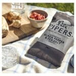 A bag of pipers black pepper & sea salt crisps on a sunny picnic blanket with bread, a bowl of beans, and glasses of water.