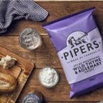 A case of Pipers Atlas Mountains Wild Thyme & Rosemary Sharing Crisps 15x150g on a wooden table, surrounded by a glass of water, bread, dip, and a striped napkin.