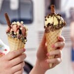Two hands holding elaborately topped ice cream cones with Dr. Oetker Chocolate Chips White 750g in front of a food truck, with a blurred figure standing in the background.