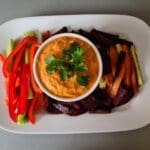 A bowl of Hummus dip garnished with cilantro, surrounded by sliced red peppers, carrots, cucumbers, and roasted beets on a white platter.