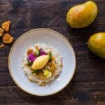 A plate of risotto made with Tilda Arborio Risotto Rice 5kg, accompanied by mango slices and edible flowers, alongside whole mangoes and bread pieces on a wooden table.