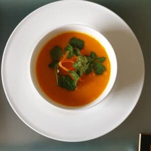 A bowl of creamy carrot soup garnished with fresh cilantro leaves, viewed from above on a white plate.