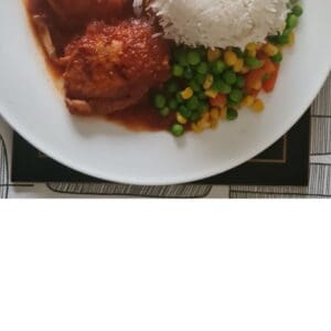 A plate of homemade food featuring two pieces of tomato chicken curry, white rice, and a mix of peas, carrots, and corn.