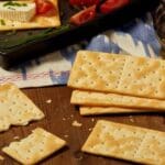 Schar Cream Crackers 210g scattered on a wooden table with cheese, tomatoes, chives, and a bottle of olive oil in the background.
