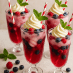 A tall dessert glass filled with creamy vanilla soft-serve made from Arla Slower Melt Soft Ice Mix, sparkling pink berry soda, and layers of fresh raspberries, strawberries, and blueberries. Finished with a swirl of ice cream and a mint sprig, this refreshing British summer dessert drink is beautifully styled on a bright wooden table. Recipe and image by Nicol Retailer Limited.