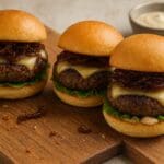 Gourmet gluten-free beef sliders with caramelised onions on rustic wooden board – recipe and image created by Nicol Retailer.