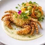Golden crispy tempura soft shell crab served with yuzu kosho emulsion, pickled cucumber and carrot, and micro herbs on a fine dining plate